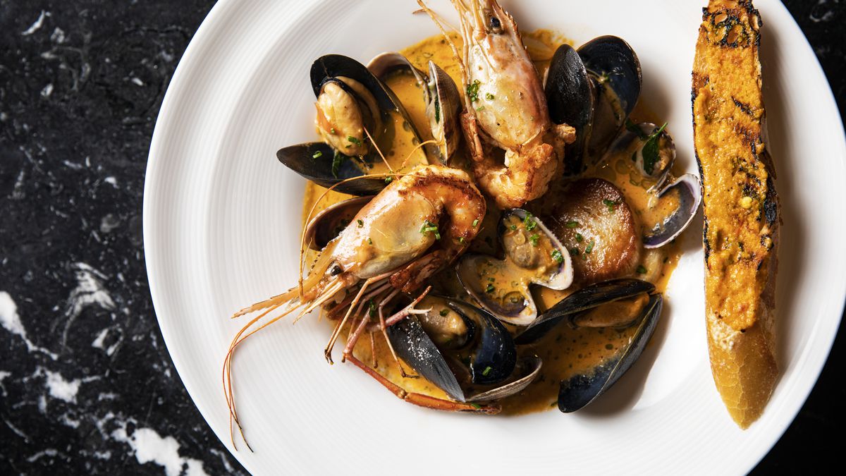 A white plate on a black-and-white granite countertop with large scallops, whole shell-on prawns, and mussels in an orange sauce.