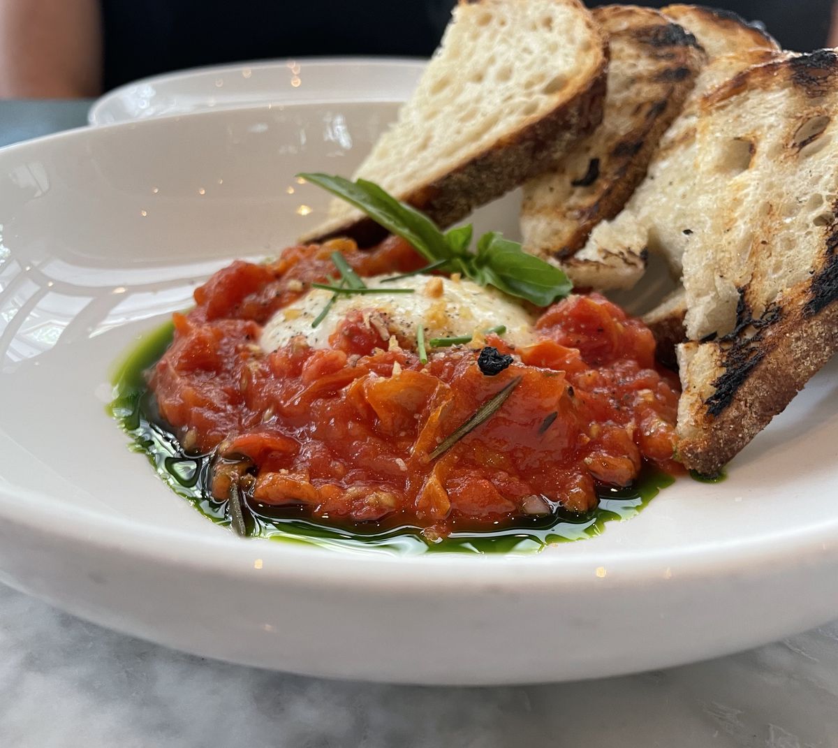 A serving of burrata is nestled into a tomato jam, that sits atop basil infused olive oil. Slices of grilled sourdough bread line the dish.