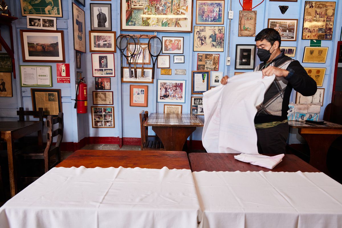 A server shakes out a tablecloth before spreading it over a table. The room is decorated with old images and objects. 