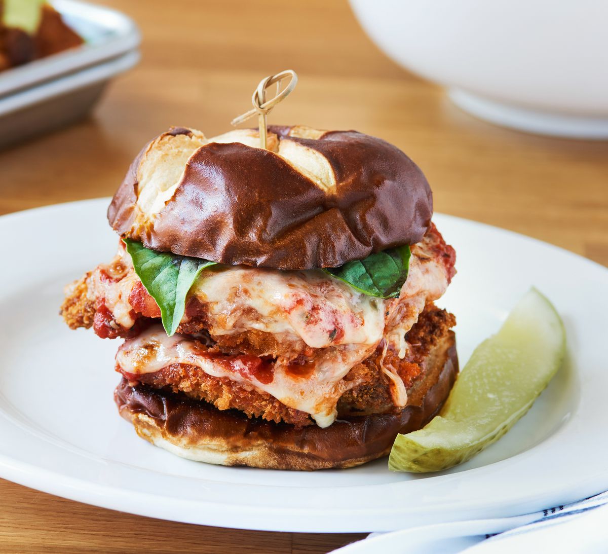A toasted hamburger bun with two pieces of battered and fried crispy chicken slathered with melted mozzarella cheese and red sauce. The sandwich is topped with bright green basil and served with a pickle spear