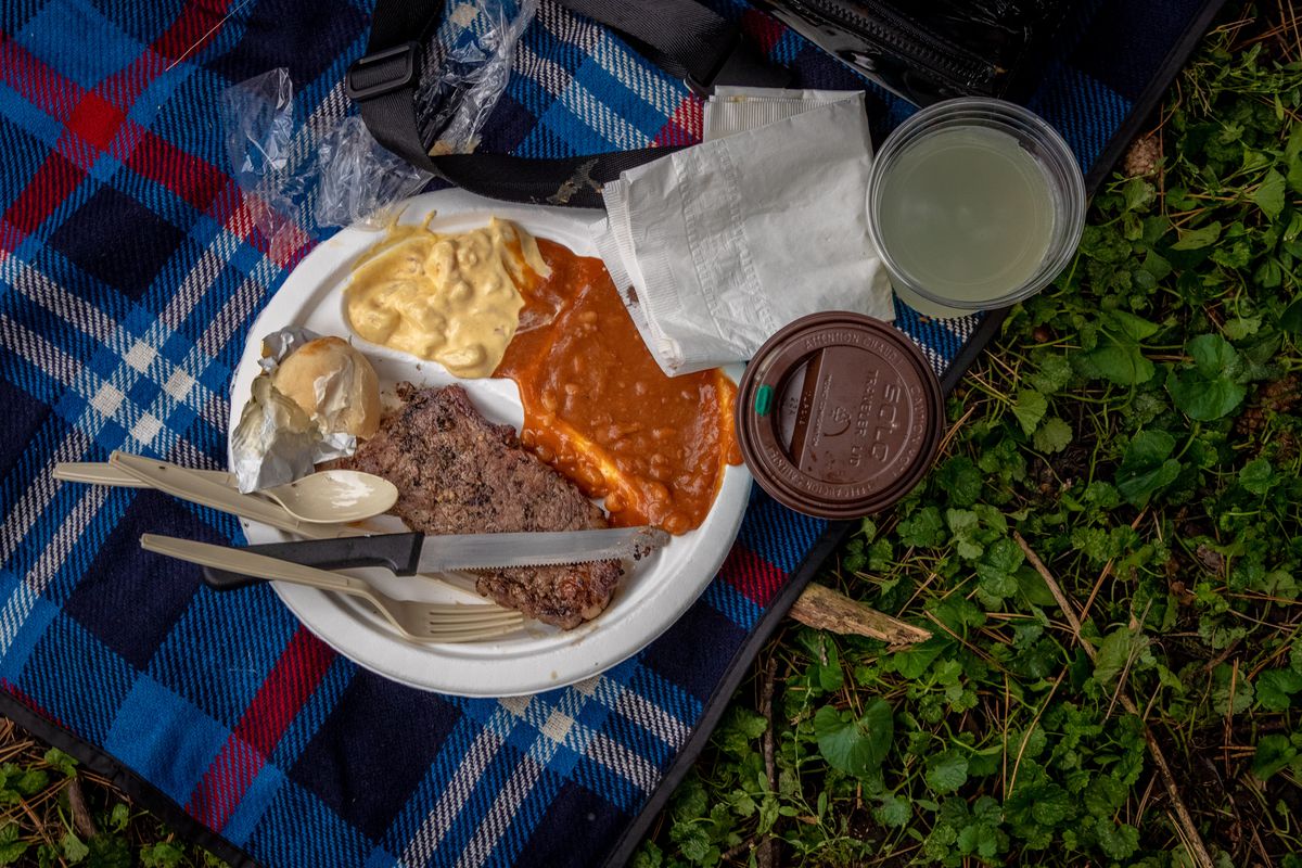 A paper plate with steak, macaroni and cheese, baked beans, and utensils sits on the grass next to a disposable cup of coffee and glass of lemonade.