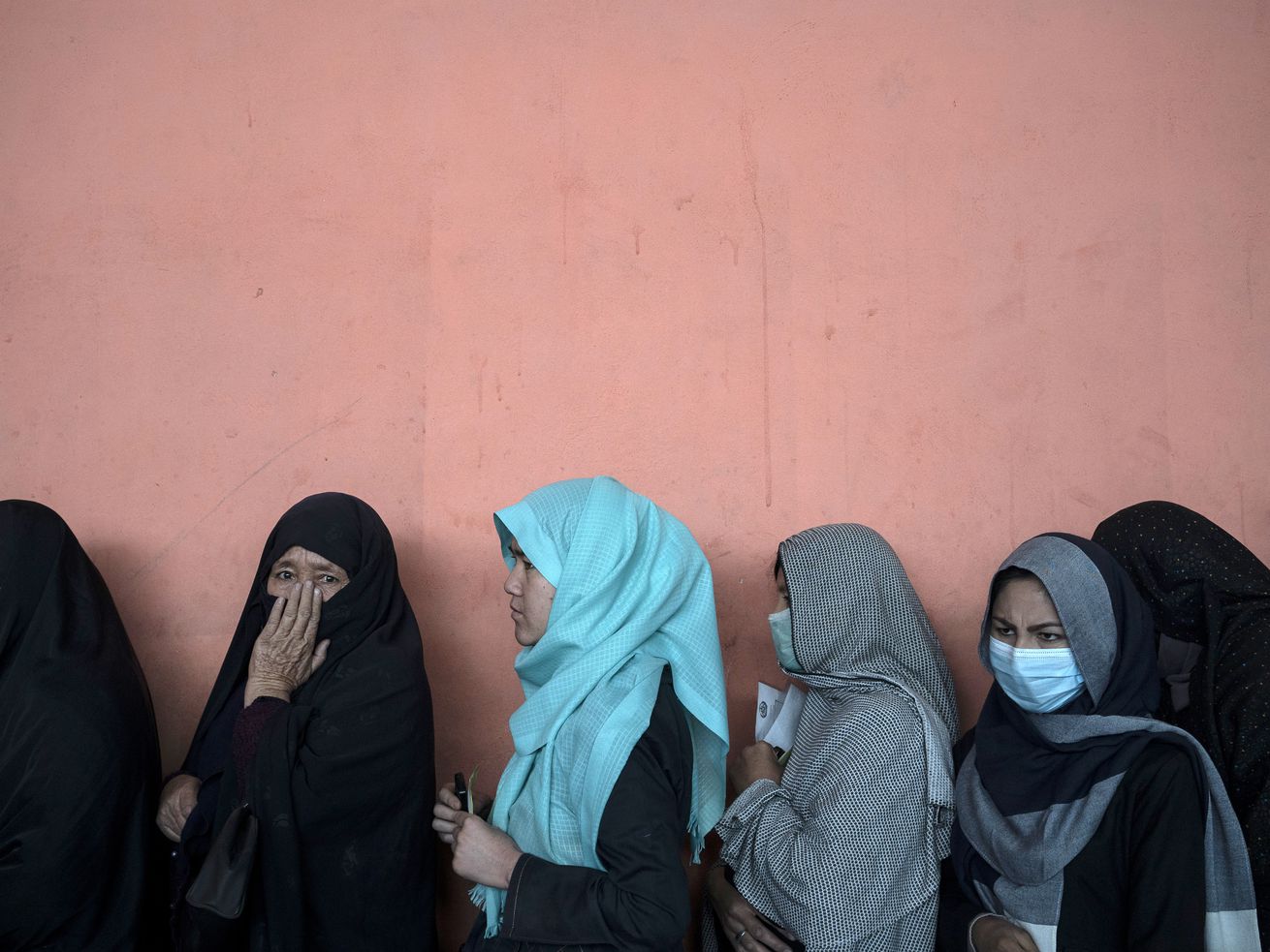 Several women wearing headscarves stand in a line against a blank wall.