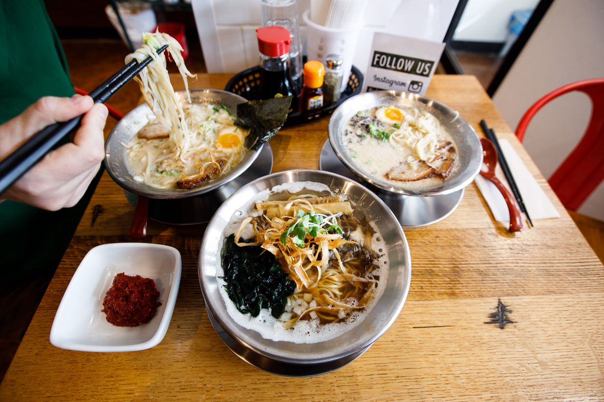 Three bowls of ramen on a wooden table with a woman’s hands holding black chopsticks that are pulling up ramen noodles. 