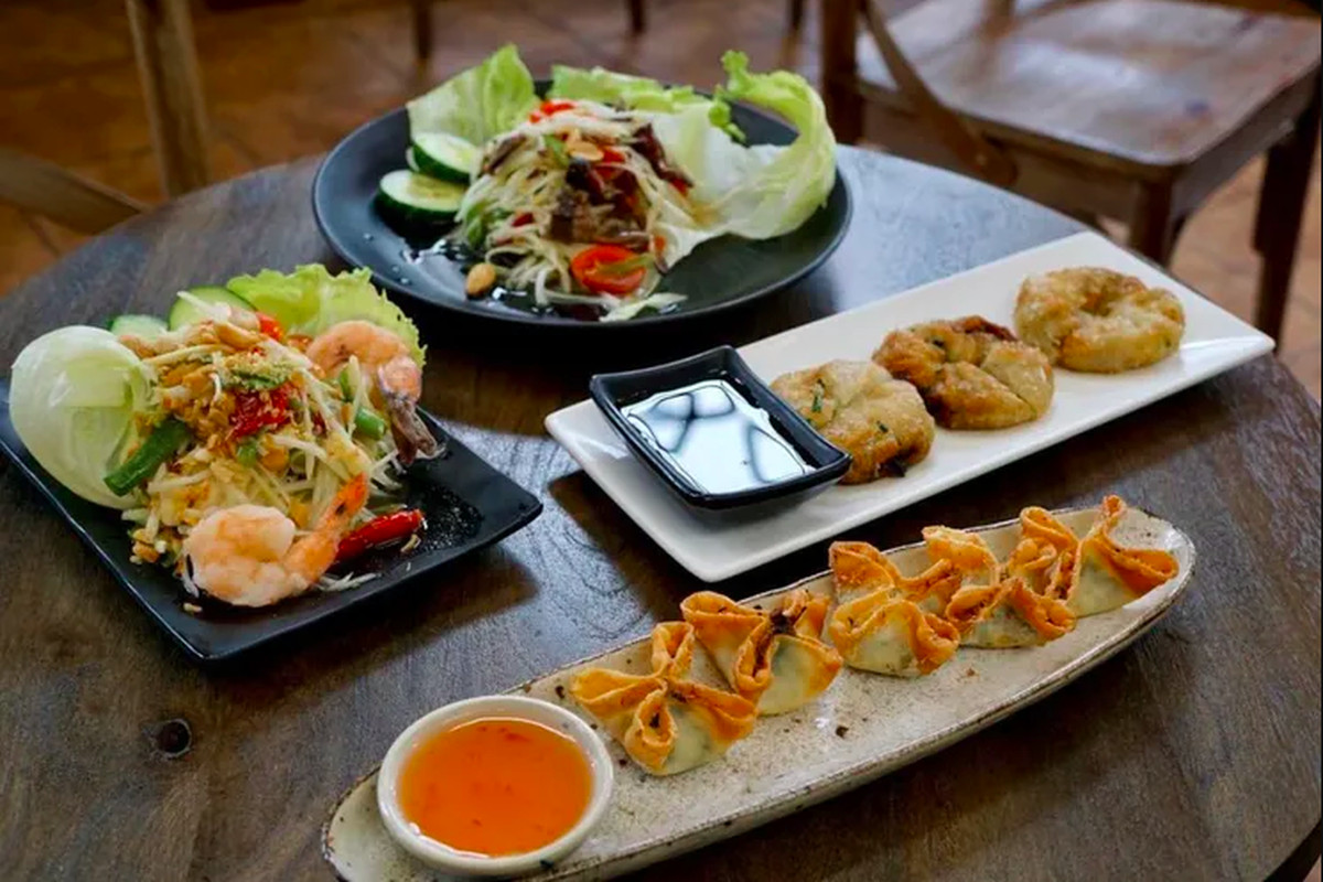 Four dishes sit on a table: in the front, wontons line up on an oblong plate, with a shrimp salad, greens, and fried disc-shaped appetizers on the other plates