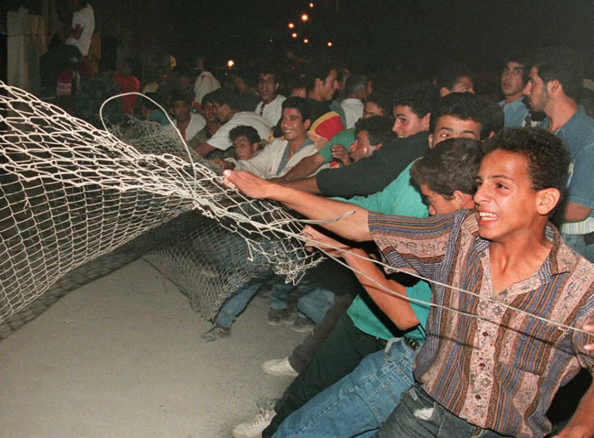 At night, and illuminated by the camera’s flash, a large group of young Palestinian men dressed in colorful ’90s clothes pull down a chain link fence. Many smile and laugh; there seems to be a festive atmosphere.