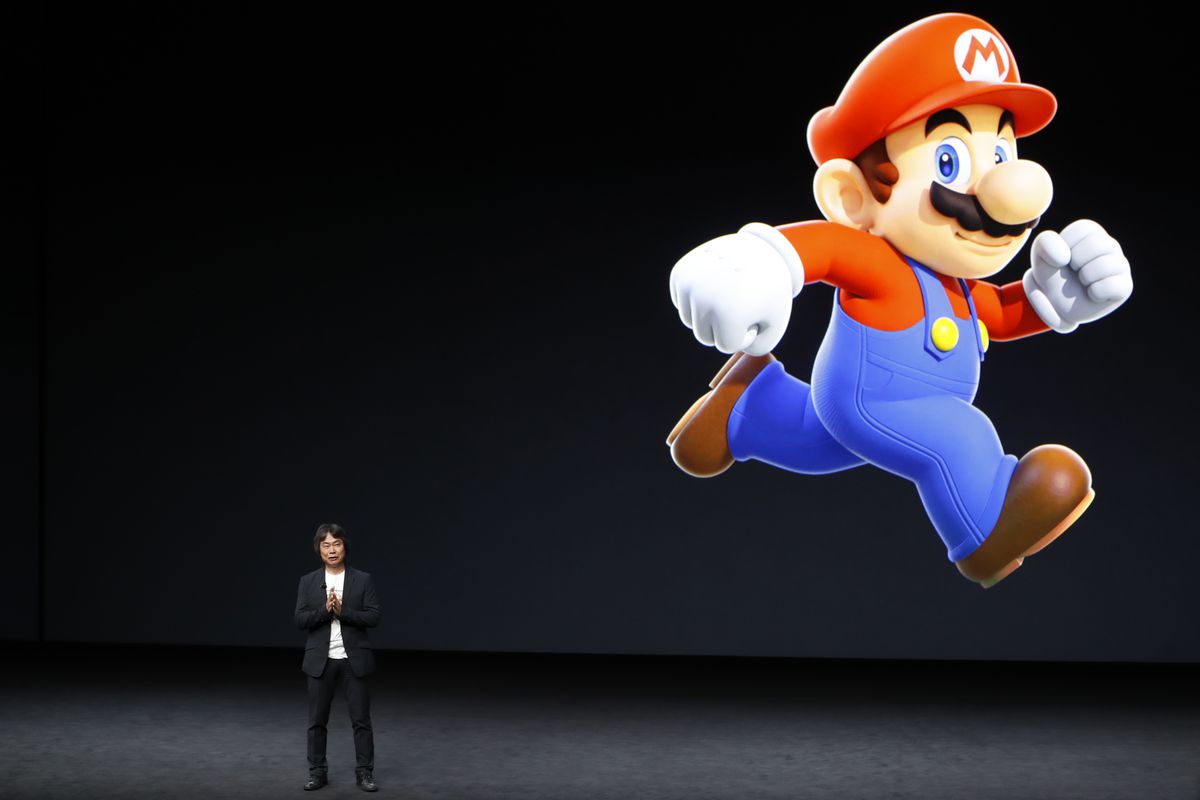 Shigeru Miyamoto speaks on a stage with a giant Mario running in the backround