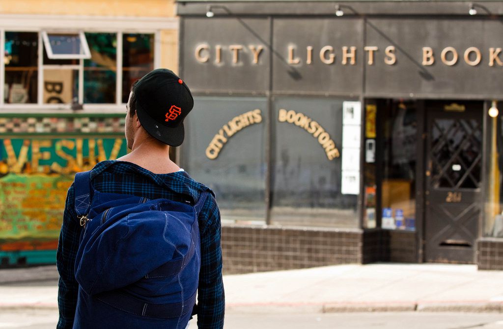 In the foreground is a person wearing a blue plaid shirt with a large blue backpack on their back. In the distance are various businesses. One of the storefronts has a sign that reads: City Lights Bookstore.