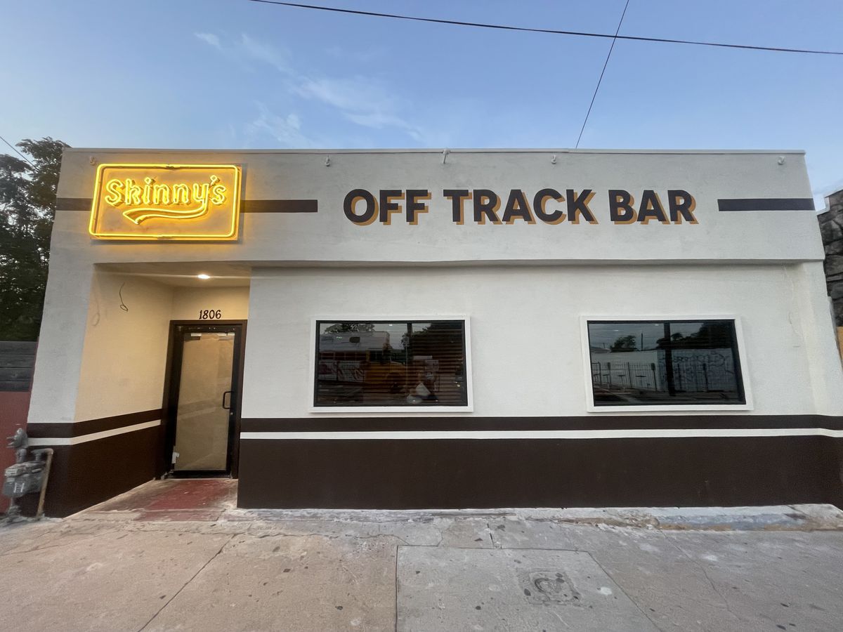 A bar facade with a yellow neon sign reading “Skinny’s” and the words Off Track Bar painted onto the white wall.