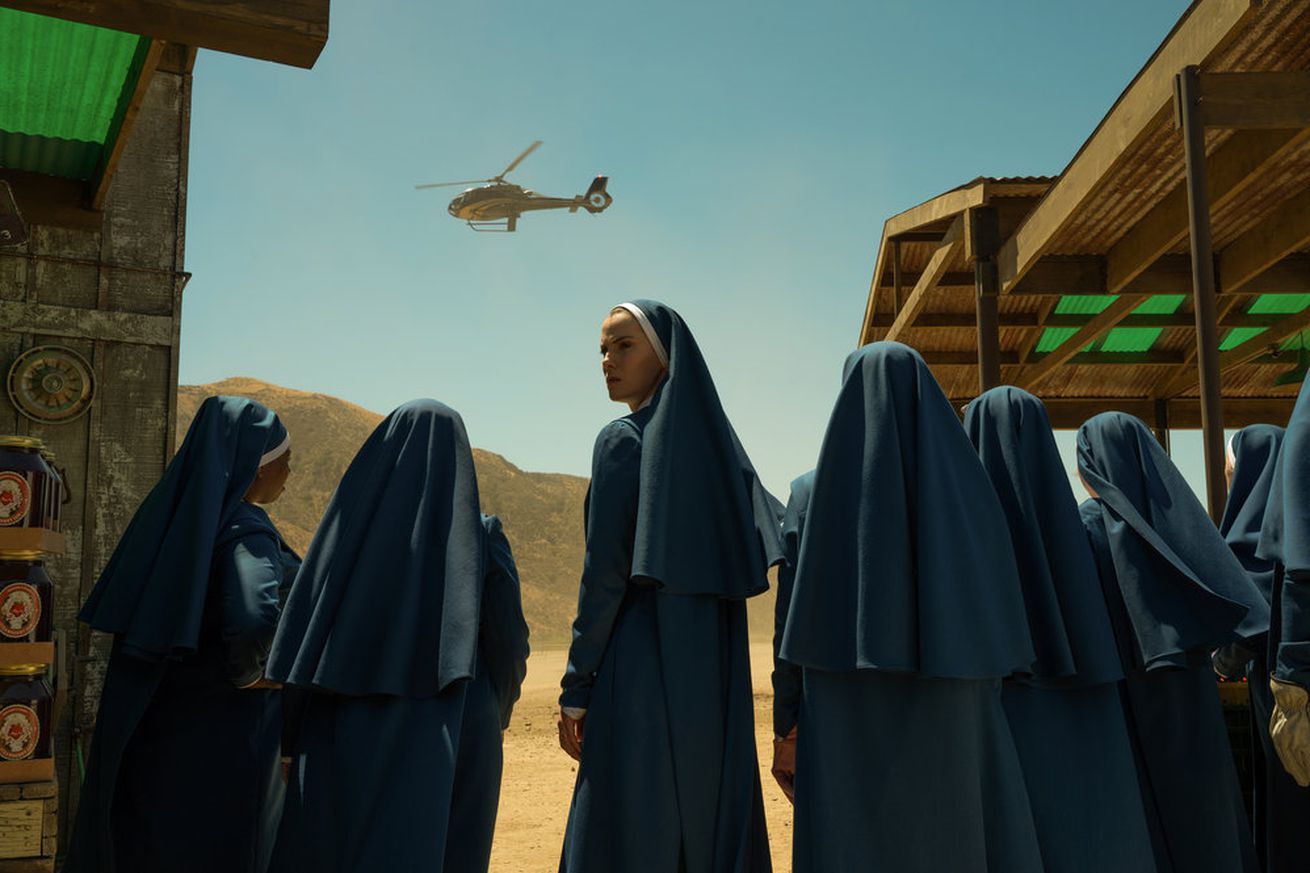 An image of multiple women dressed in blue nun robes, standing in the desert, watching a helicopter land. One nun looks back, concerned.