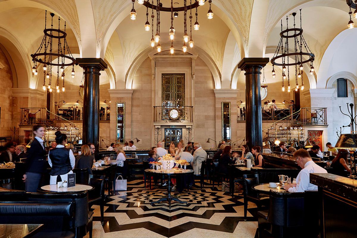 The dining room at the Wolseley in London, with a star-pattern tiled floor, black chandeliers hanging pendant bulbs, and two waiters standing attention.