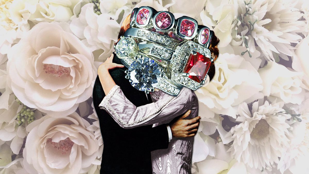A couple kisses in front of a floral background with a stack of diamond and ruby rings in front of their faces