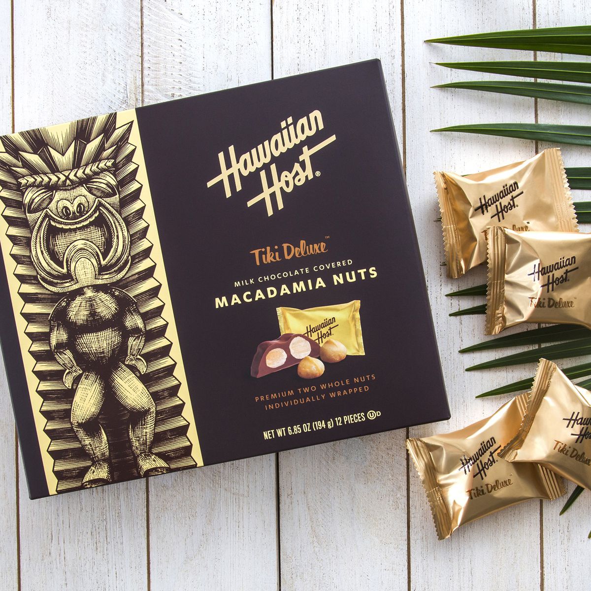 A box of Hawaiian Host Tiki Deluxe Milk Chocolate Macadamia Nuts, sitting on a deck next to a palm leaf and several wrapped candies.