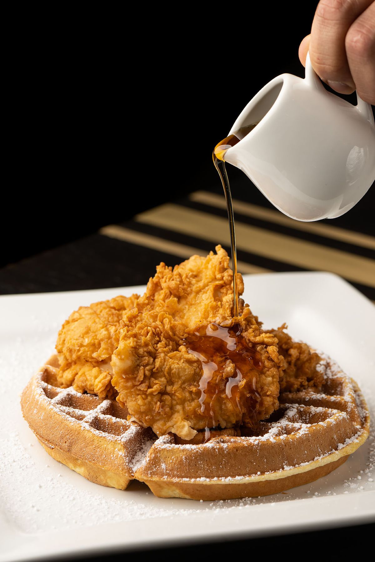 An overhead pour of syrup onto fried chicken and a waffle below.