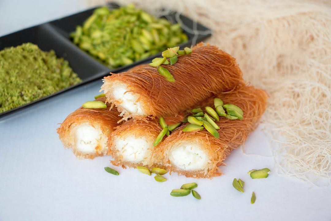 Rolls of knafeh stacked on a white surface, with piles of ingredients in the background