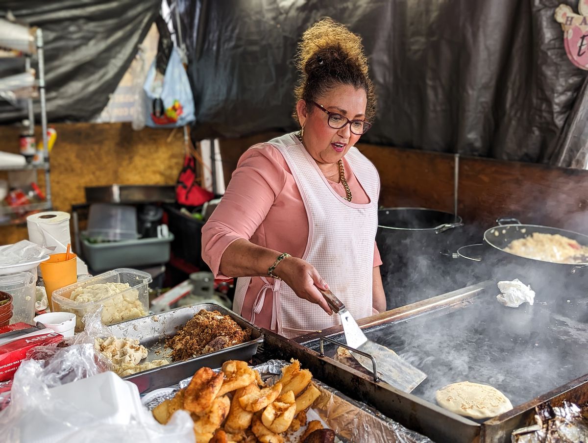 Woman wearing a pink dress and apron cooking pupusas.