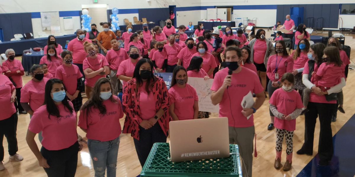 A school gym full of parents and children wearing masks and red t-shirts stare at a laptop where they were participating in a virtual meeting.