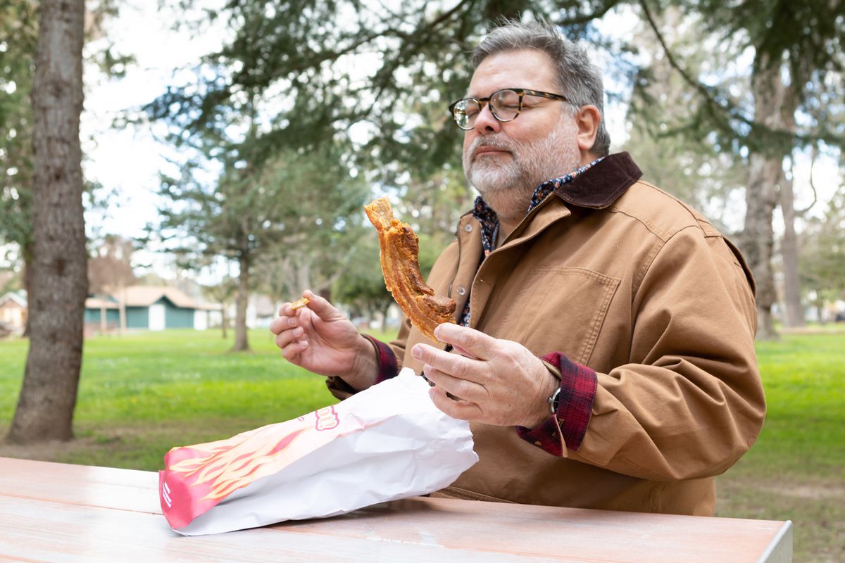 Steve Sando sits at a picnic table holding a large piece of chicharrones.
