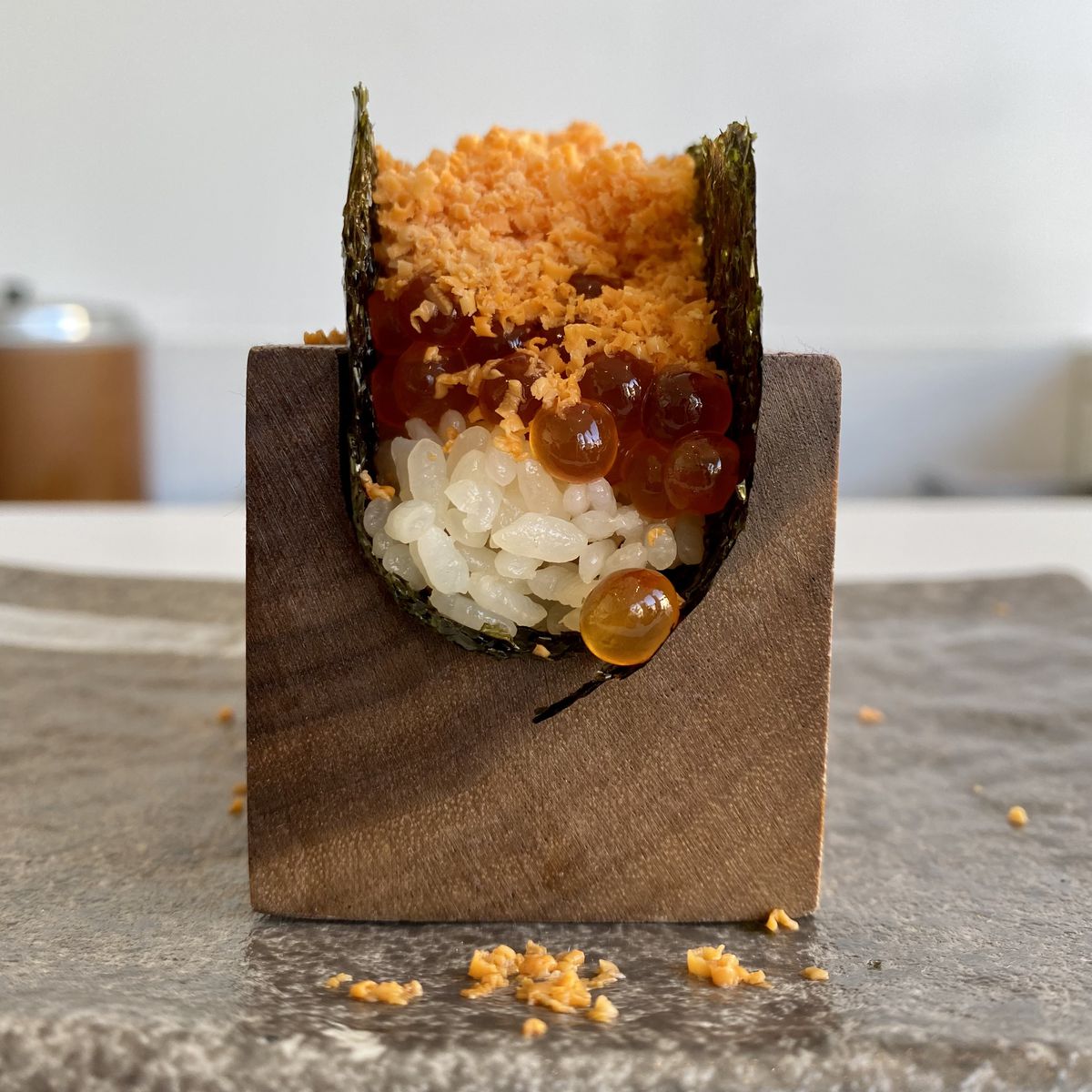 A handroll made with rice, ikura, and grated front monkfish liver held in a wooden U-shaped stand on a counter.
