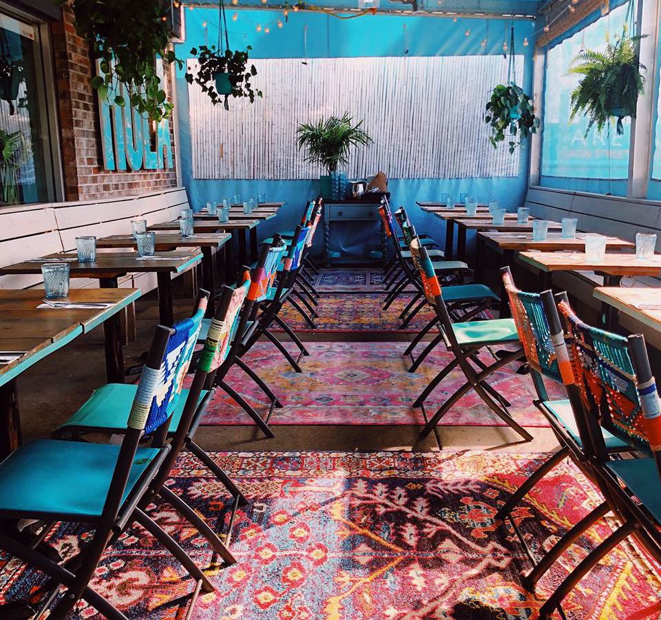 A tented patio with pattered rugs on the floor and Hola’s signature turquoise colored chairs at the tables