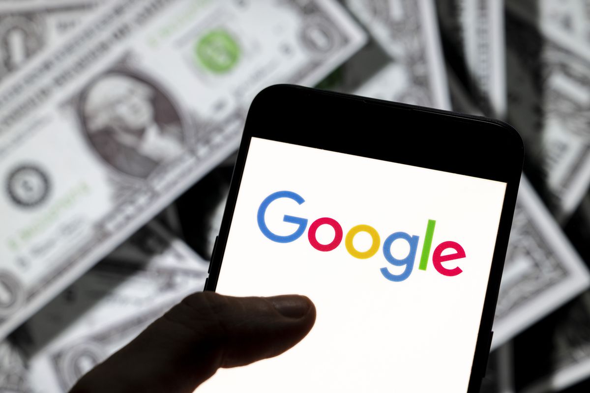 A photo illustration shows a pile of dollar bills behind a hand holding a smartphone screen displaying the Google logo.