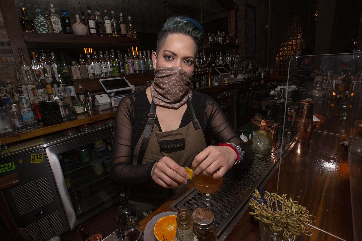 A woman in a mask stands behind a bar garnishing a cocktail.