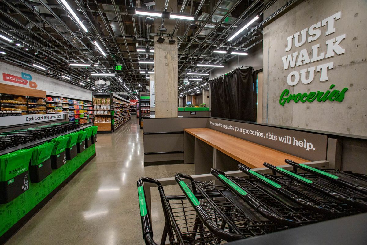 A sign says “Just Walk Out” next to shopping carts and a wooden counter to bag groceries.