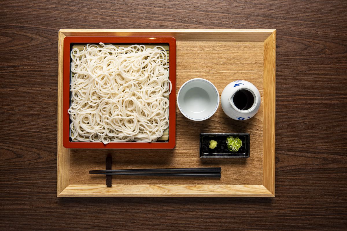 An overhead photograph of a red tray of white noodles, arranged beside a tray of wasabi, ginger, and dipping sauce.