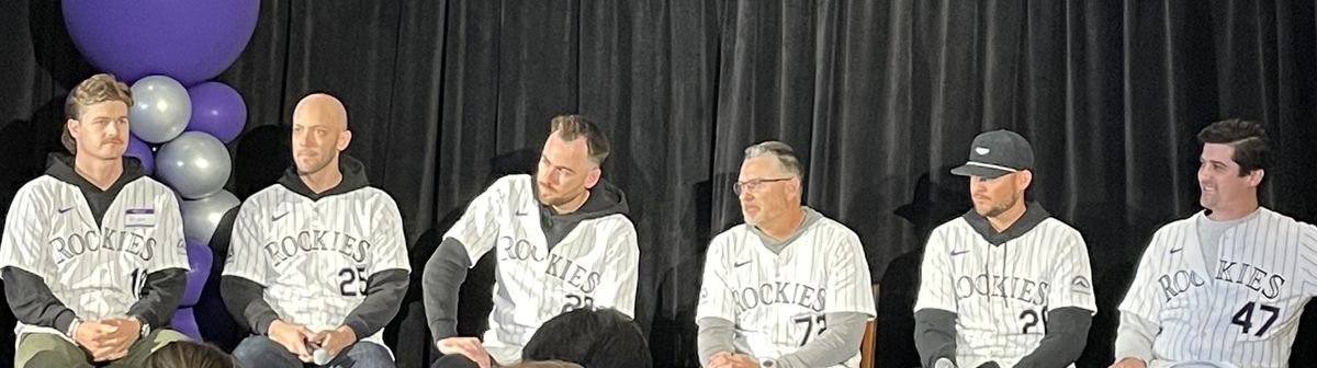 Picture of four Rockies pitchers, one catcher, and the pitching coach sitting on stools in their uniforms with a black curtain behind them. 