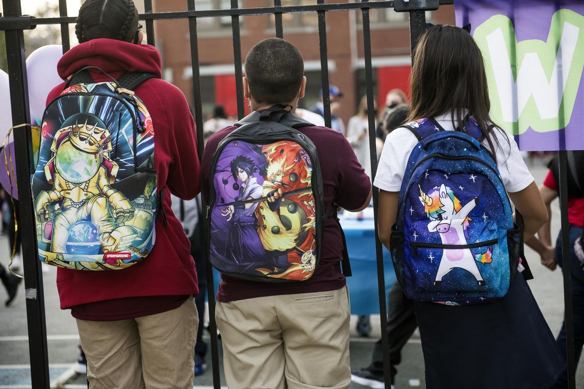 Three students wearing backpacks look through a fence. Their faces aren’t visible.