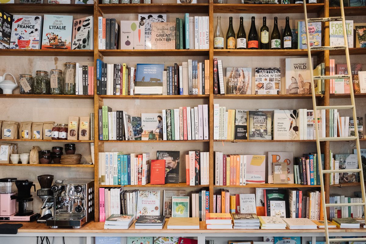 Books, wine bottles, an espresso maker, and bags of coffee line tall bookshelves, with a ladder resting on one end.