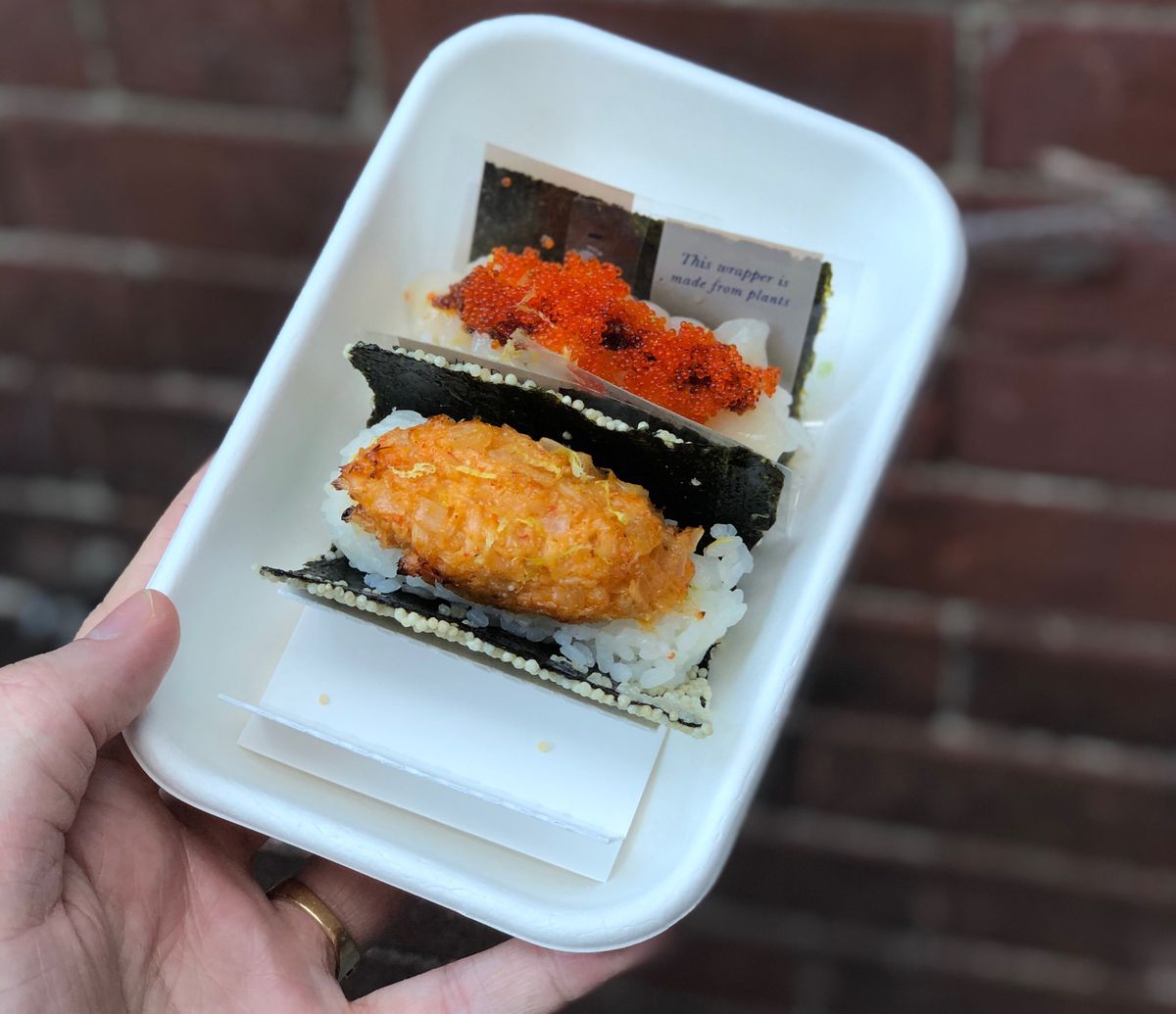 Two sushi rolls in a white platter held up by a hand against a red-brick background