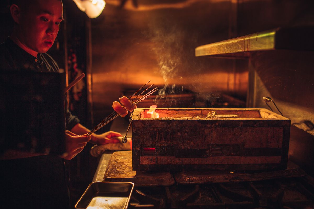Under a glowing orange light in an industrial kitchen, a bald Taiwanese chef holds two pieces of fish on a small skewer, about to put it over an open flame in in a wooden box.