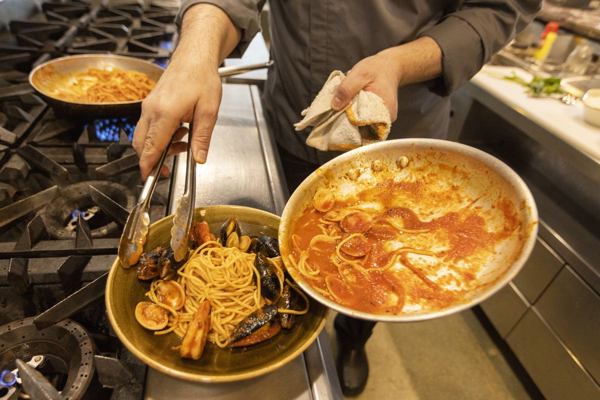 A chef uses tongs to move a pasta dish from a skillet to a bowl.