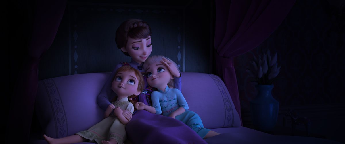 Queen Iduna, a pale brunette woman, cuddles with young Elsa and Anna on their bed
