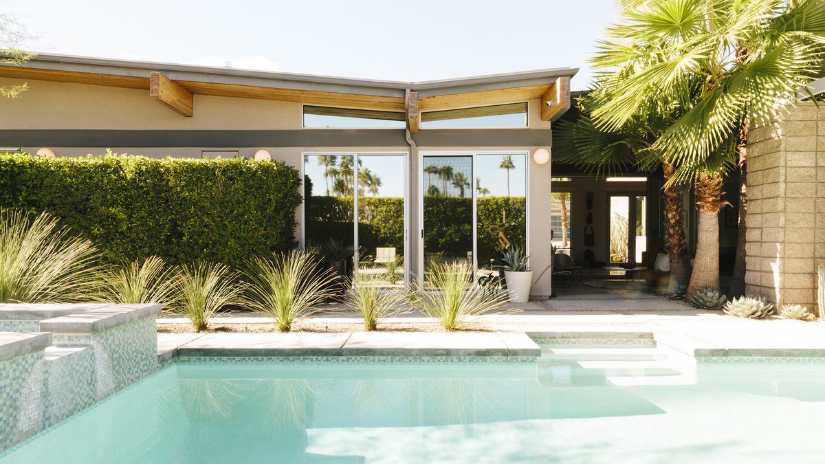 A swimming pool with light blue water is in the foreground. There is a house in the background with glass doors. A palm tree and other plants sit between the house and the swimming pool. 