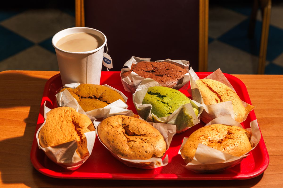 A red tray has seven sponge cakes in varying shades of brown with a cup of coffee with milk on it.