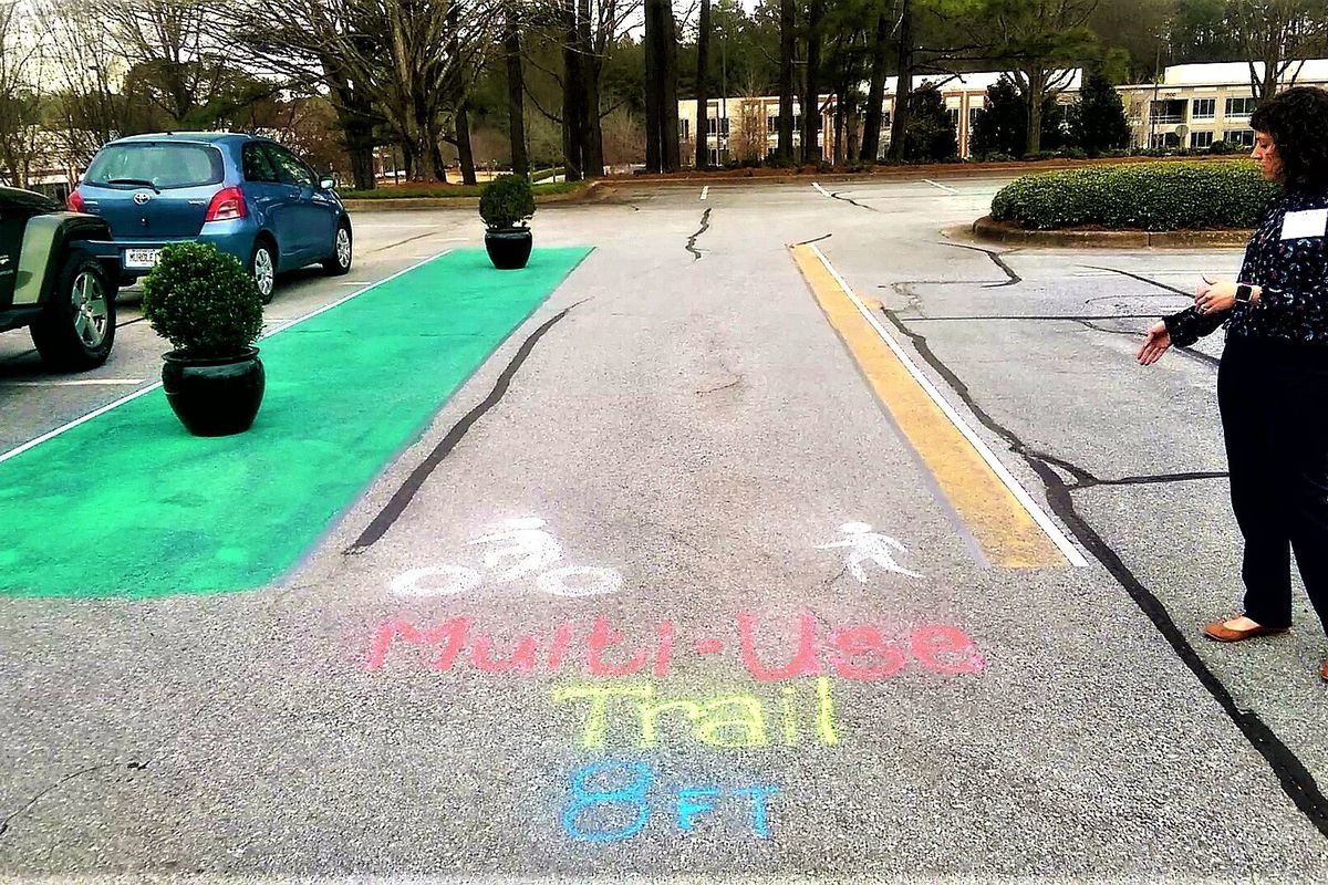 A woman in a parking lot points to a mocked-up version of a multi-use trail drawn with chalk and paint.