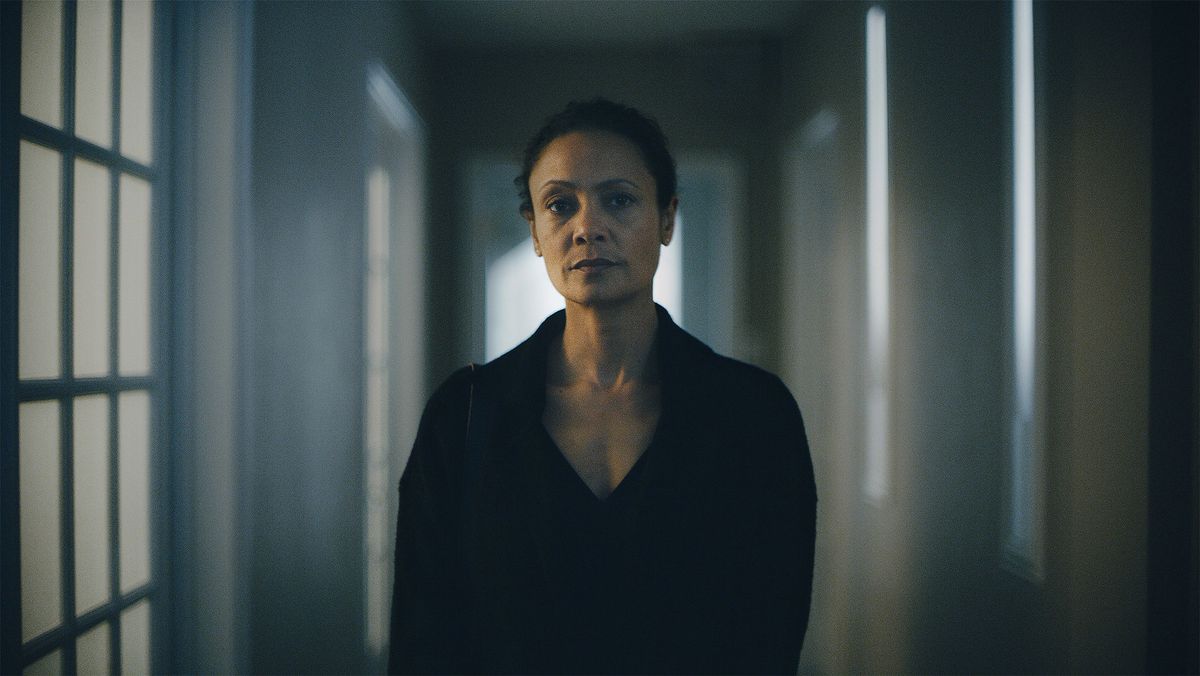 A black woman stands alone in the center of the frame. She’s in a hallway, and there are shadows on the walls.