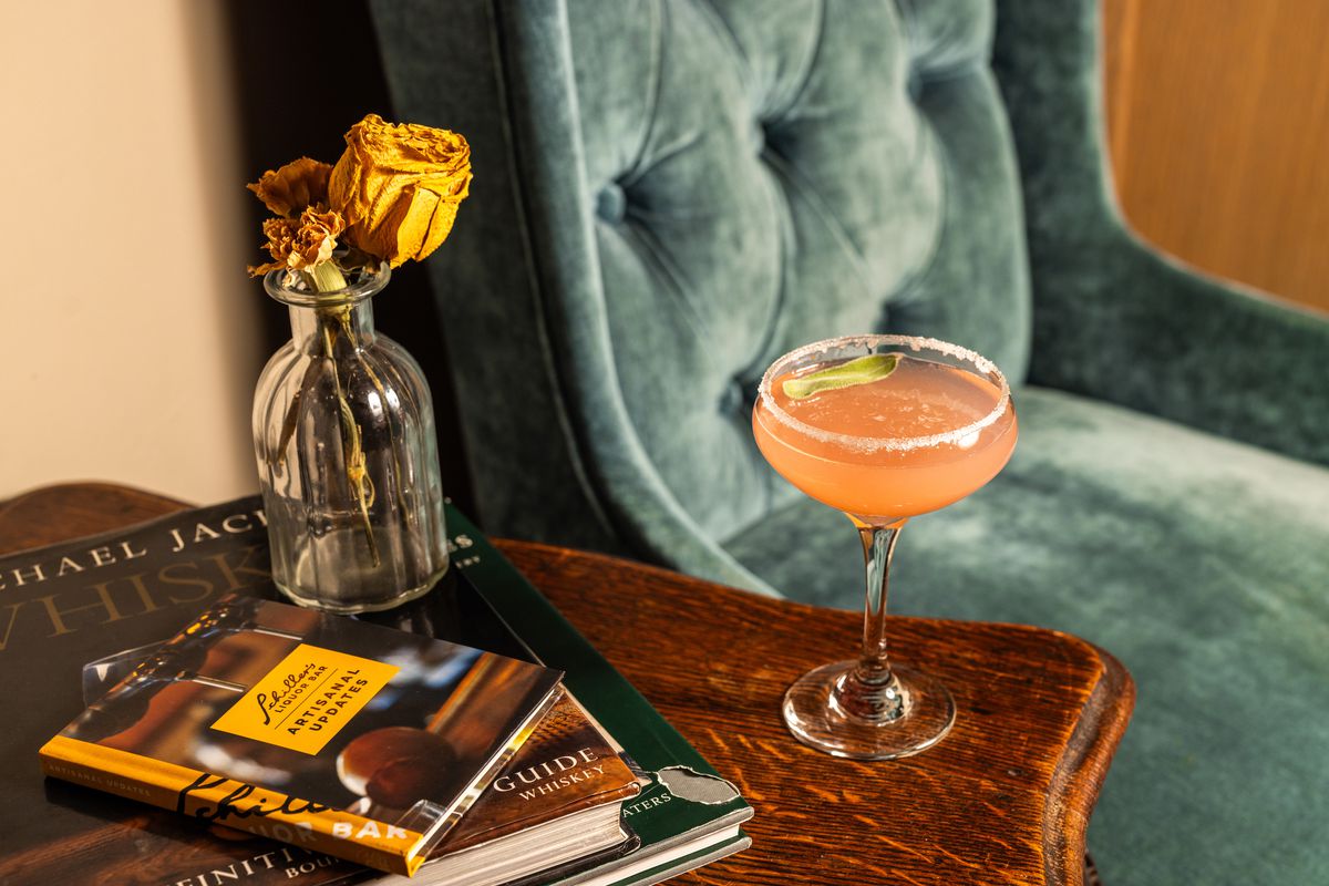 A pink cocktail is served in a coupe. It sits on an end table with a vase of dried flowers and several guide books. Next to it is a plush blue chair.
