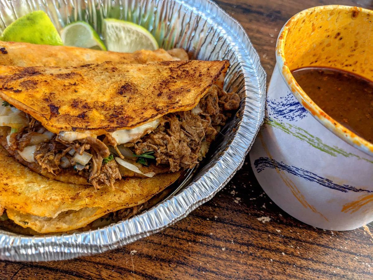 Three birria tacos in browned shells sit in a circular aluminum takeout container with lime wedges. A paper cup of reddish broth sits to the side.