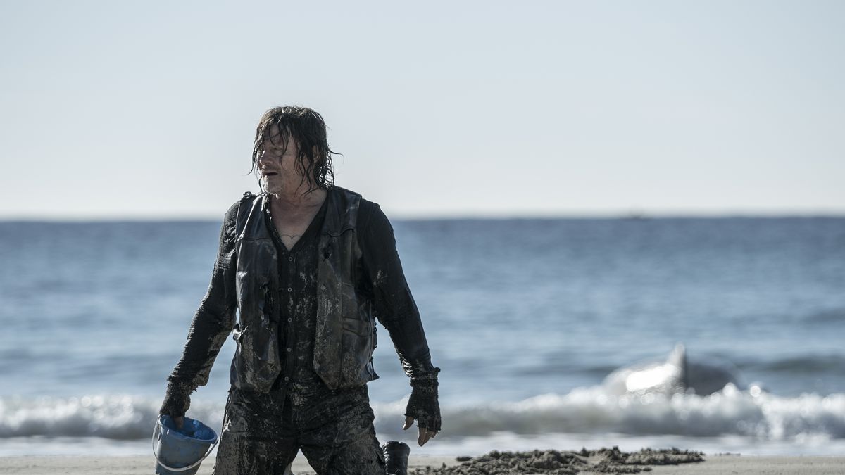 Daryl Dixon (Norman Reedus) kneeling on a beach with a bucket looking wet and worn
