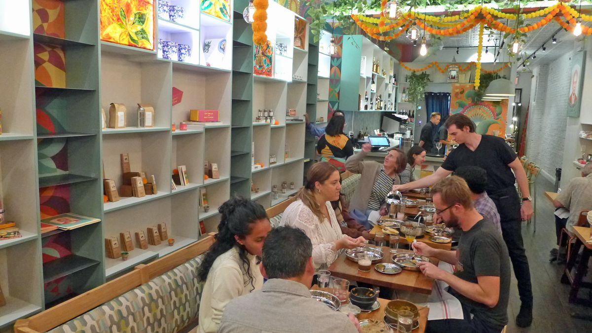 A room with shelves of groceries on the left and diners sitting at tables in the middle, as swags of artificial flowers dangle from the ceiling.