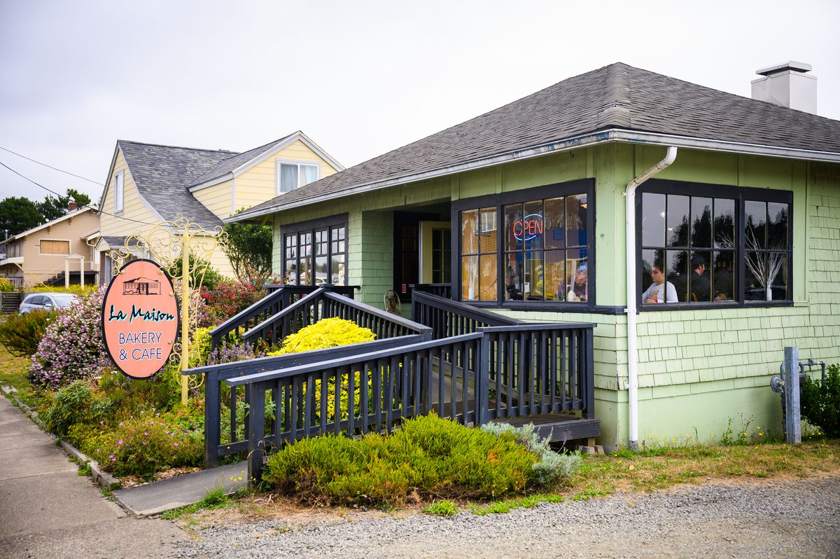 A green house surrounded by bushes in Newport, Oregon. An orange sign reads “La Maison Bakery &amp; Cafe.”