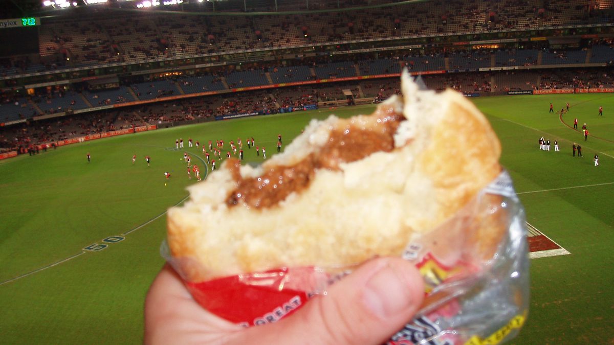 A hand holds a meat pie in a sports stadium