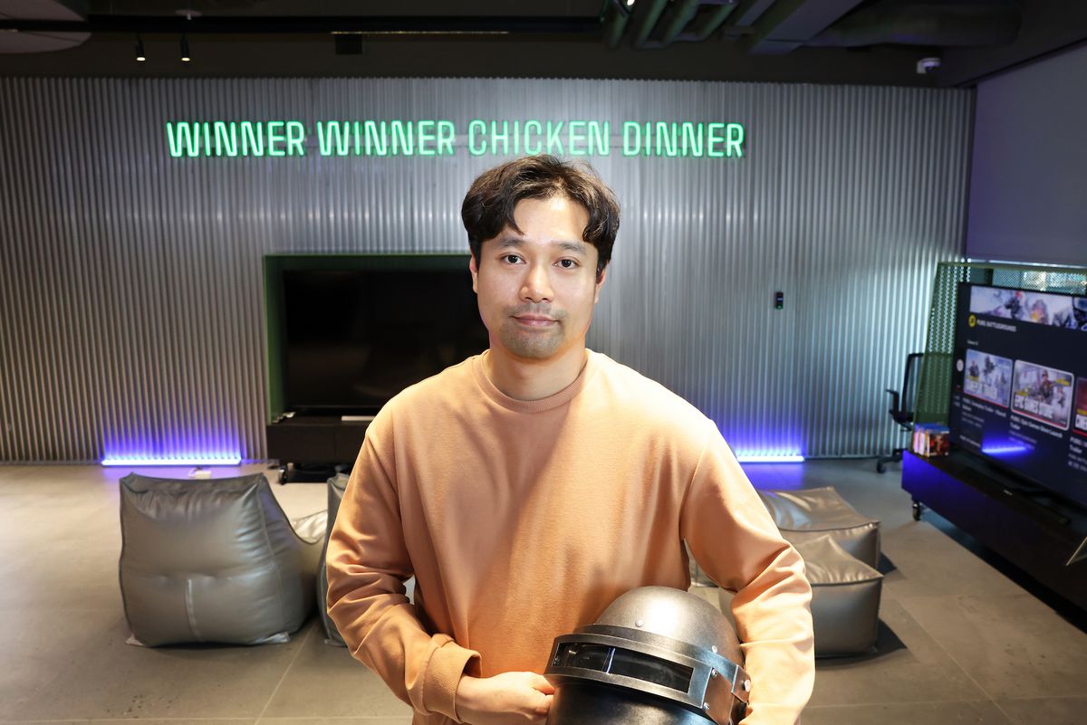 A photograph of Taeyhun Kim of PUBG Studios holding the classic PUBG helmet and standing in front of a neon sign that reads “Winner winner chicken dinner.”
