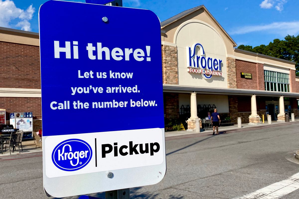 A blue and white sign advertising curb-side grocery pickup in a parking lot in front of a Kroger’s grocery store on a sunny day.