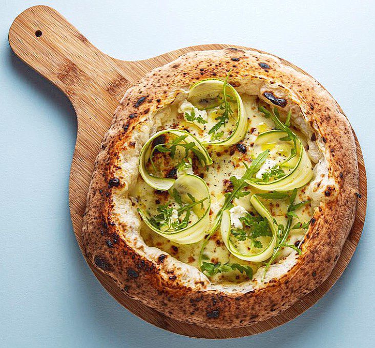 A full pizza with a thick fluffy crust on a wooden pizza paddle on a neutral background. The white pizza is topped with shaved zucchini