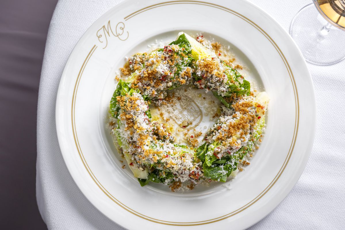 A white plate embossed with a gold rim and the initials “MC” sits on a white tablecloth. It holds a gem lettuce Caesar salad arranged in a circle and sprinkled with dressing and breadcrumbs.