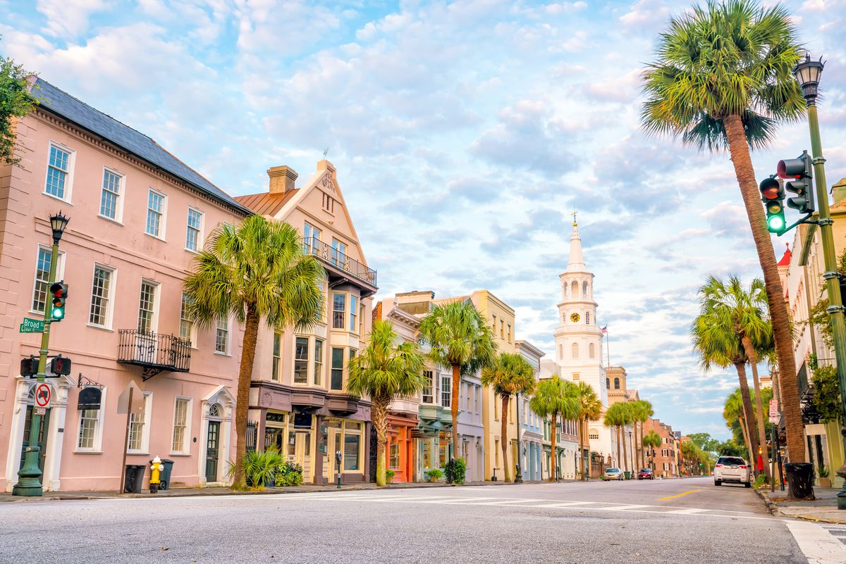 A view of a street in downtown historic Charleston. The buildings are pastel colored and there are palm trees lining the sidewalks.