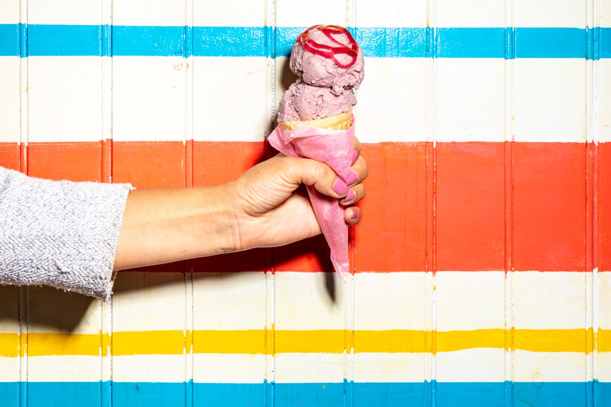 A hand with pink nails holds up an api morada ice cream cone for a photograph against a white, blue, and yellow striped wall